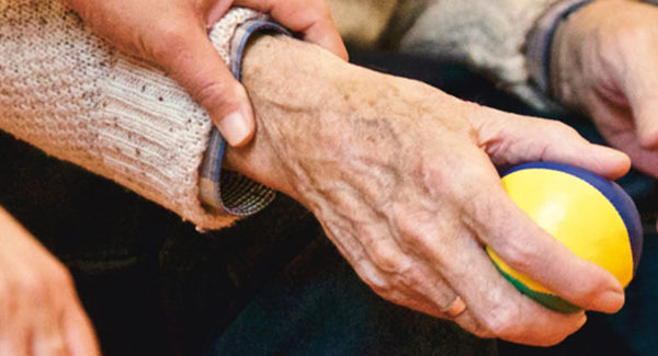 An older person's hand gripping a tennis ball. A caregiver is holding the hand.
